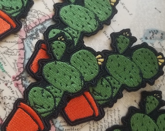 May include: A set of three embroidered cactus patches featuring green cacti and orange pots. The patches are displayed on a vintage map background.