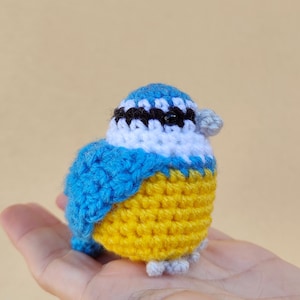 Może przedstawiać: Ręcznie robiony, szydełkowy ptak w kolorze niebieskim z żółtym brzuszkiem, białym piersią i czarnymi oczami. Ptak siedzi na dłoni.