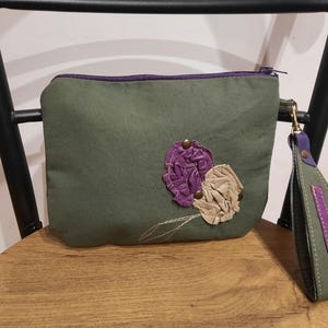 May include: An olive green pouch with a zip closure and wrist strap. The pouch is decorated with purple and beige fabric flower appliqués and embroidered details. The wrist strap is olive green with purple accents.