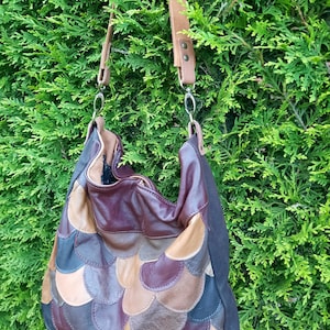 Op de afbeelding: Een hobo-tas met een veelkleurig patchwork-ontwerp van overlappende lederen schubben in tinten bordeaux, beige, bruin en donkerblauw. De tas is voorzien van een beige lederen schouderband bevestigd met zilverkleurige metalen clips.
