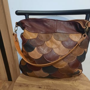 Può includere: Una borsa a spalla in stile hobo realizzata con toppe di pelle sovrapposte in varie tonalità di marrone, beige e grigio scuro. La borsa presenta un motivo a squame, un pannello superiore in pelle marrone scuro e una tracolla in pelle chiara.