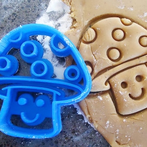 Puede incluir: Un cortador de galletas de plástico azul con forma de hongo con una cara sonriente. El cortador tiene una superficie texturizada con pequeños círculos en la parte superior del hongo.