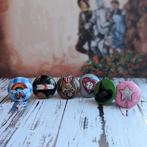 May include: A collection of seven decorative, round cabinet knobs. Each knob features a unique design, including a rainbow, a scroll, a medal, a heart, a witch's hat, and a star. The knobs are painted in various colors and set against a blurred background.