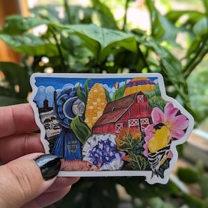 May include: A sticker in the shape of Iowa, featuring a red barn, corn, a blue ribbon, and a yellow bird. The sticker has the words "Iowa State Fair" on it. The background is a blue sky with a hot air balloon.