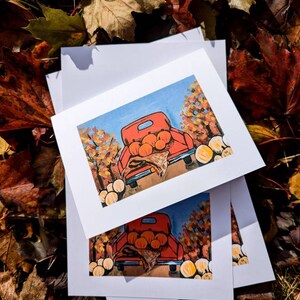 May include: Three white cards with a red truck carrying pumpkins in a fall setting. The truck is surrounded by trees with orange, yellow, and red leaves. The cards are stacked on top of each other.