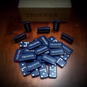 May include: A set of blue dominoes with white dots and the word "TROOPER" printed on each tile. The dominoes are arranged in a pile on a wooden surface.