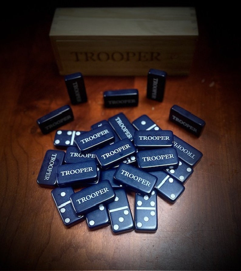 May include: A set of blue dominoes with white dots and the word "TROOPER" printed on each tile. The dominoes are arranged in a pile on a wooden surface.