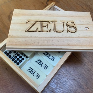 May include: A wooden box with the word "ZEUS" engraved on the lid. The box contains a set of dominoes with the word "ZEUS" printed on them.