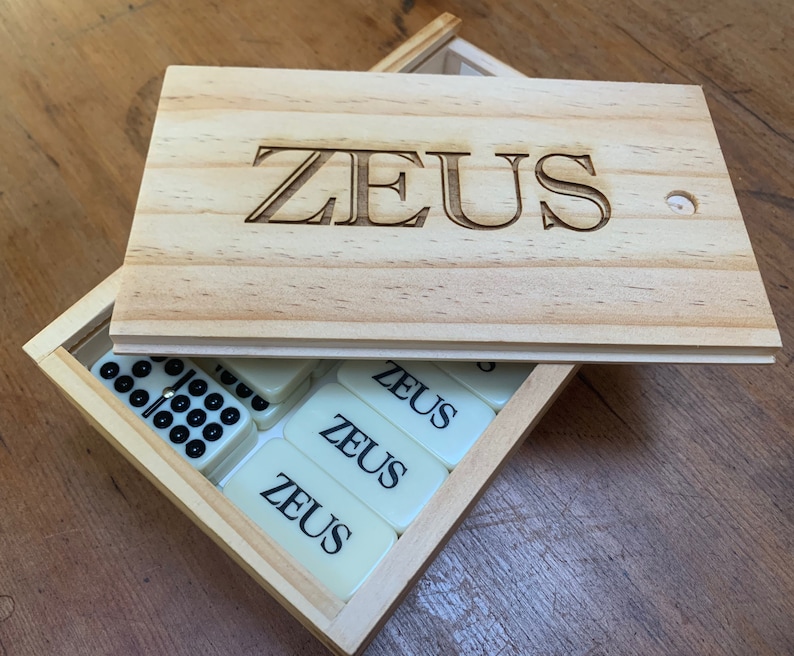 May include: A wooden box with the word "ZEUS" engraved on the lid. The box contains a set of dominoes with the word "ZEUS" printed on them.
