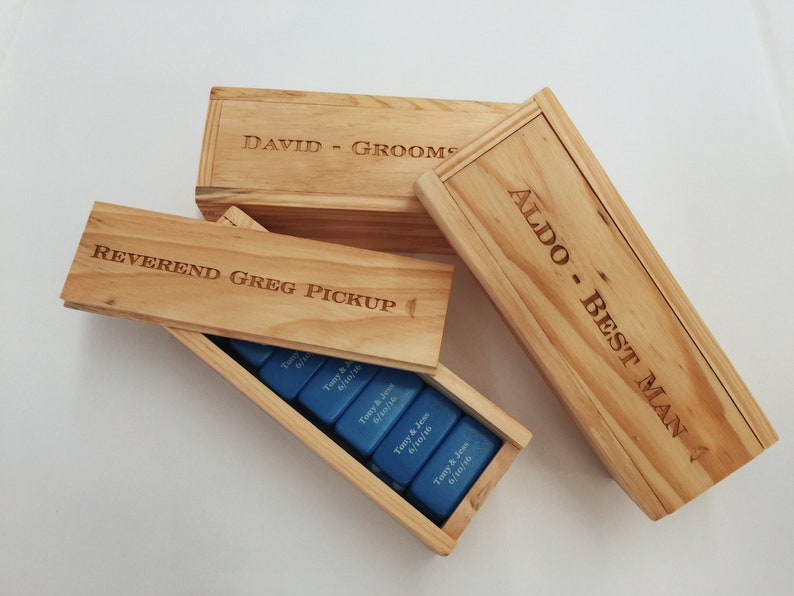 May include: Three wooden boxes with engraved names and titles: "David - Groom", "Reverend Greg Pickup", and "Aldo - Best Man". The boxes are stacked on top of each other and contain blue dominoes.