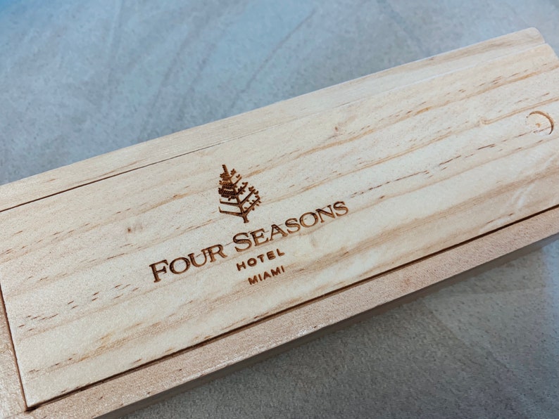 May include: A wooden box with the Four Seasons Hotel Miami logo engraved on the top. The logo features a stylized tree and the text "Four Seasons Hotel Miami".