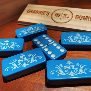 May include: A set of blue dominoes with white floral designs and the initials "SL" on each tile. The dominoes are arranged on a wooden surface with a sign that reads "Grannie's SL Dominoes".