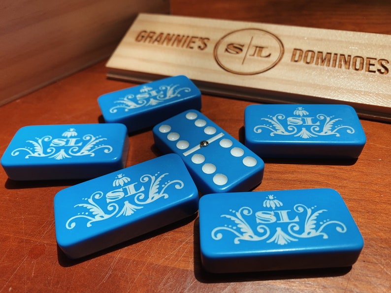 May include: A set of blue dominoes with white floral designs and the initials "SL" on each tile. The dominoes are arranged on a wooden surface with a sign that reads "Grannie's SL Dominoes".