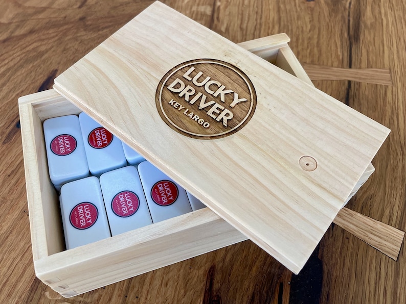 May include: A wooden box with a hinged lid, containing a set of dominoes. The lid is engraved with the words "Lucky Driver Key Largo" in a circle.