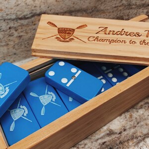 May include: A wooden box with a blue and white domino set inside. The box has a personalized inscription that reads "Andres Pacia Champion to the last stroke". The dominoes have a blue background with white dots and a logo of a rowing team.