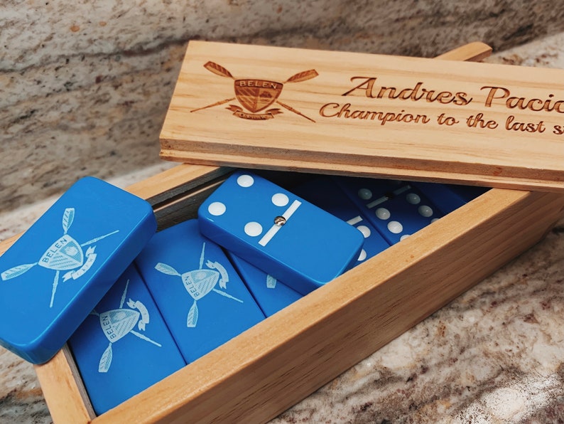 May include: A wooden box with a blue and white domino set inside. The box has a personalized inscription that reads "Andres Pacia Champion to the last stroke". The dominoes have a blue background with white dots and a logo of a rowing team.