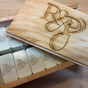 May include: A wooden box with a lid that has the initials "R & D" engraved on it. The box is filled with white dominoes that also have the initials "R & D" engraved on them.