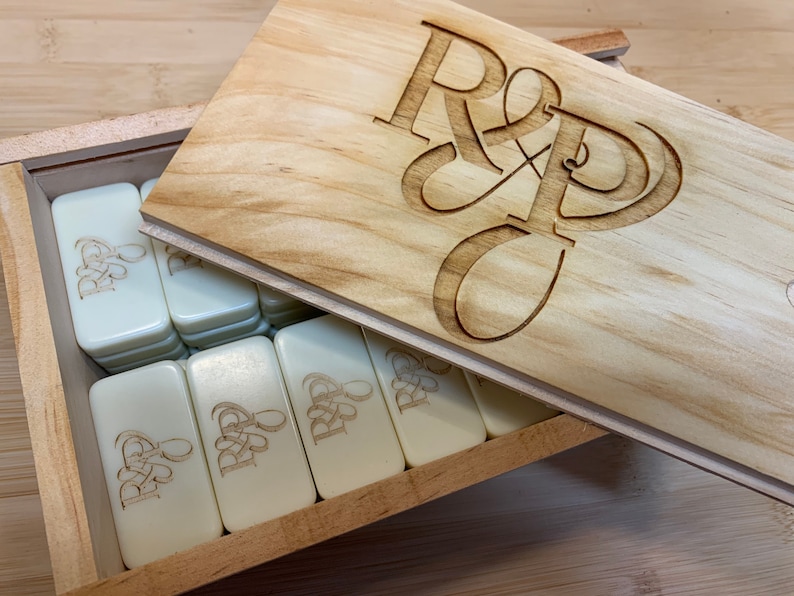 May include: A wooden box with a lid that has the initials "R & D" engraved on it. The box is filled with white dominoes that also have the initials "R & D" engraved on them.