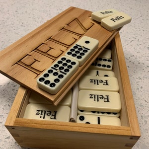 May include: A wooden box with a hinged lid, containing a set of dominoes. The lid is open and the dominoes are visible. The dominoes are white with black dots and have the word "Feliz" written on them. The box is made of light wood and has the word "Feliz" carved into the lid.