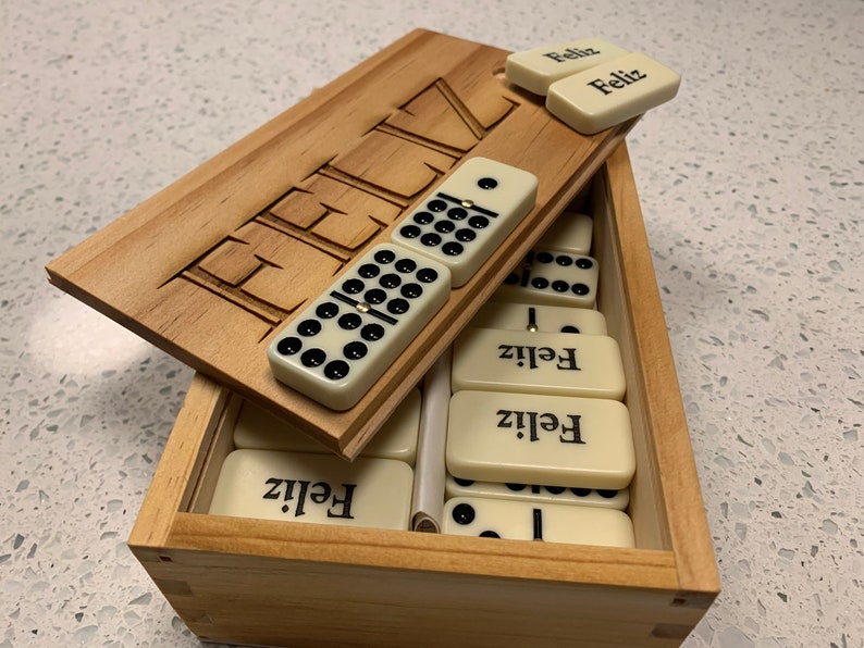 May include: A wooden box with a hinged lid, containing a set of dominoes. The lid is open and the dominoes are visible. The dominoes are white with black dots and have the word "Feliz" written on them. The box is made of light wood and has the word "Feliz" carved into the lid.