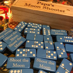 May include: A set of blue dominoes with white dots and the text "Shoot the Moon" on each tile. The dominoes are in a wooden box labeled "Papa's Moon Shooter".