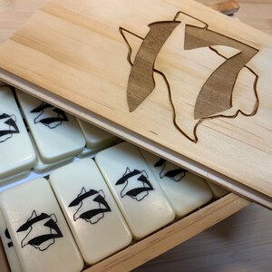 May include: A wooden box with a lid containing a set of white dominoes with a black graphic design. The lid has a carved design of the same graphic.
