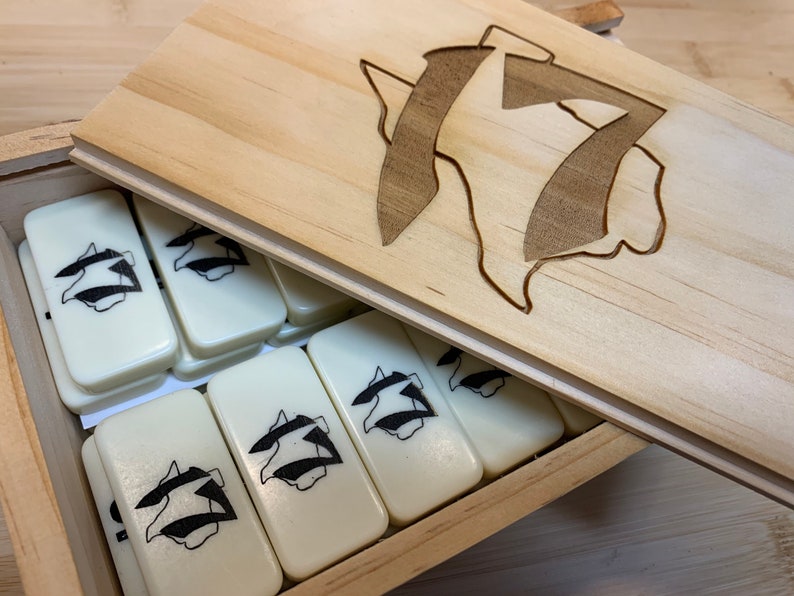 May include: A wooden box with a lid containing a set of white dominoes with a black graphic design. The lid has a carved design of the same graphic.
