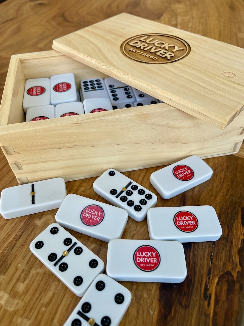 May include: A wooden box with a set of dominoes. The box is labeled "Lucky Driver Key Largo". The dominoes are white with black dots and have the same logo on them.