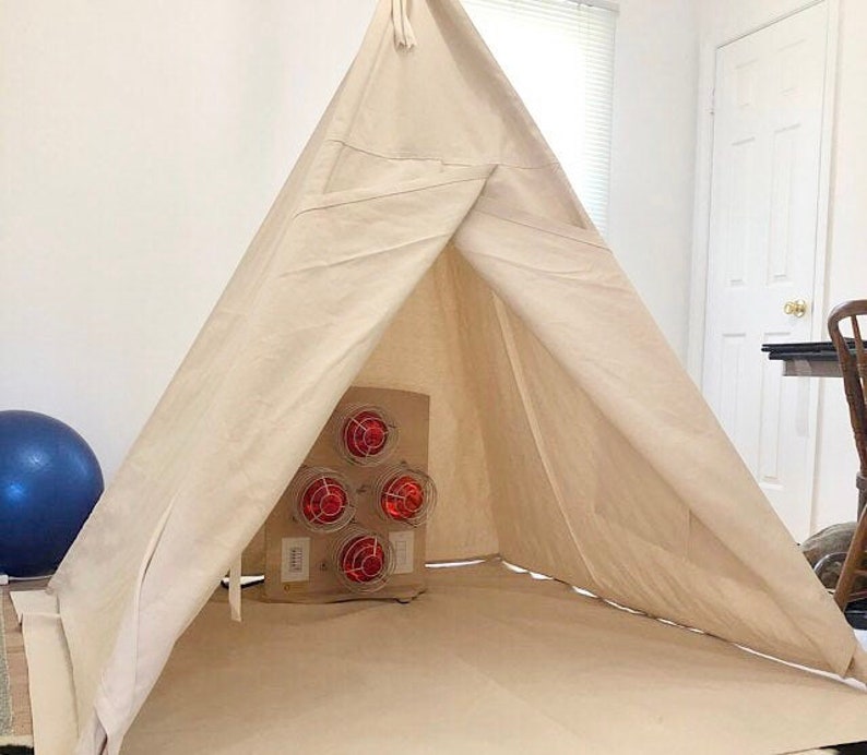 Puede incluir: Una tienda de campa&ntilde;a tipi de lona blanca con una unidad de calefacci&oacute;n roja y negra en su interior. La tienda de campa&ntilde;a est&aacute; montada sobre un suelo marr&oacute;n claro.