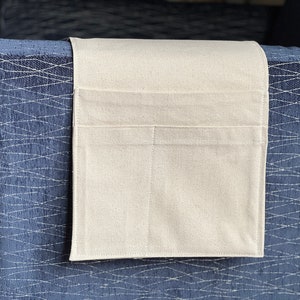Peut inclure: Un organisateur en tissu rectangulaire de couleur blanc cass&eacute; dot&eacute; de deux poches horizontales, con&ccedil;u pour &ecirc;tre suspendu sur l'accoudoir d'un meuble. L'organisateur est fabriqu&eacute; dans un textile uni avec des coutures visibles sur les bords.