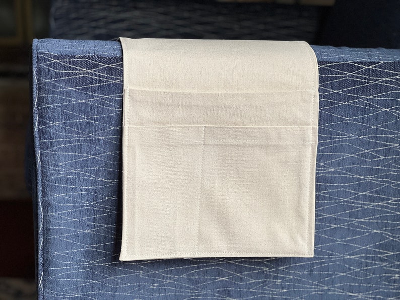 Peut inclure: Un organisateur en tissu rectangulaire de couleur blanc cass&eacute; dot&eacute; de deux poches horizontales, con&ccedil;u pour &ecirc;tre suspendu sur l'accoudoir d'un meuble. L'organisateur est fabriqu&eacute; dans un textile uni avec des coutures visibles sur les bords.