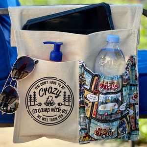 May include: A beige canvas chair organizer with multiple pockets. The organizer has a pocket with a water bottle, a pocket with a tablet, a pocket with a bottle of sunscreen, and a pocket with sunglasses. The organizer has a black and white graphic that says "You don't have to be crazy to camp with us. We will train you."