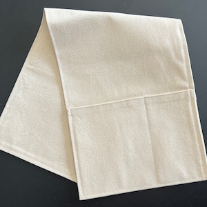 May include: A rectangular, off-white fabric runner featuring a single large pocket sewn onto the front surface. The item is constructed from a plain-weave textile with visible stitching along the edges and the pocket perimeter, creating a clean, functional design.