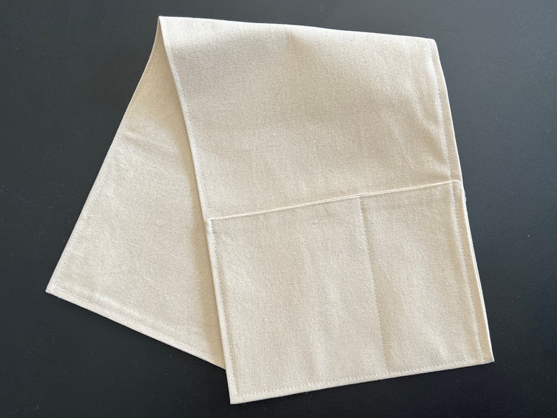 May include: A rectangular, off-white fabric runner featuring a single large pocket sewn onto the front surface. The item is constructed from a plain-weave textile with visible stitching along the edges and the pocket perimeter, creating a clean, functional design.