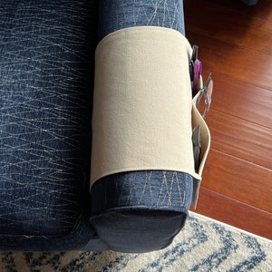 Peut inclure: Un organisateur d'accoudoir en tissu beige pos&eacute; sur l'accoudoir d'un canap&eacute; bleu fonc&eacute; textur&eacute;. L'organisateur comporte plusieurs poches lat&eacute;rales contenant des stylos et une paire de lunettes, offrant un rangement pour les petits objets.