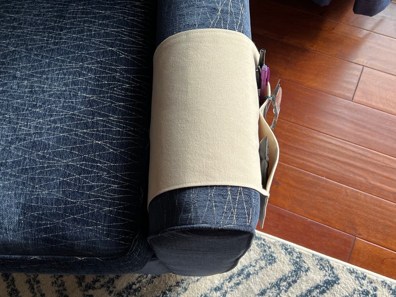 Peut inclure: Un organisateur d'accoudoir en tissu beige pos&eacute; sur l'accoudoir d'un canap&eacute; bleu fonc&eacute; textur&eacute;. L'organisateur comporte plusieurs poches lat&eacute;rales contenant des stylos et une paire de lunettes, offrant un rangement pour les petits objets.