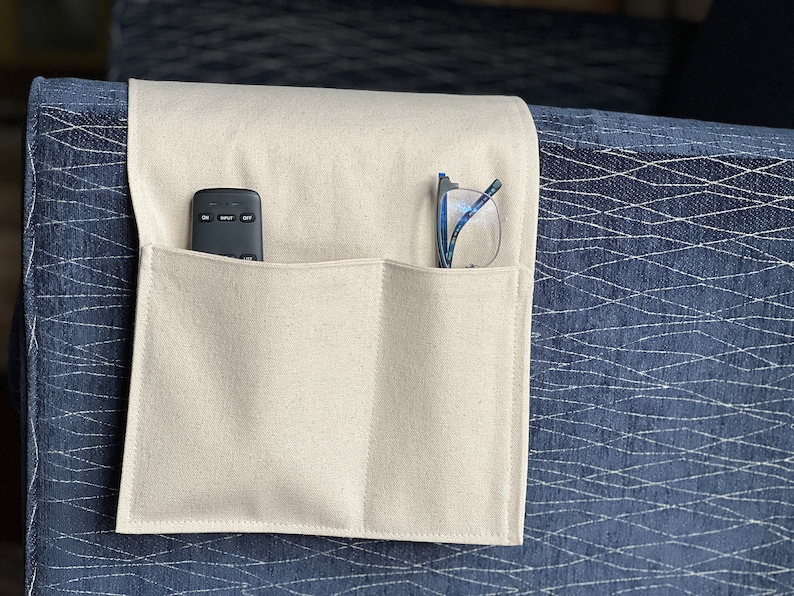 May include: A beige fabric armrest organizer featuring two rectangular pockets draped over a dark blue textured sofa arm. The left pocket contains a black remote control, and the right pocket holds a pair of eyeglasses with blue frames.