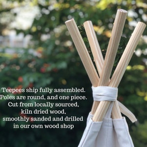 May include: Close-up of the top of a teepee tent with four wooden poles converging at the top. The poles are natural wood color and have a smooth finish. The teepee is made of white canvas fabric. The text "Teepees ship fully assembled. Poles are round, and one piece. Cut from locally sourced, kiln dried wood, smoothly sanded and drilled in our own wood shop" is visible at the bottom of the image.