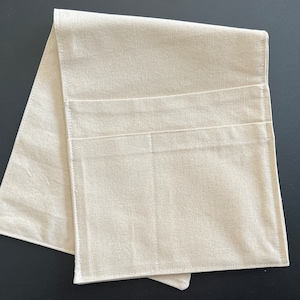 Peut inclure: Un organisateur en tissu beige clair de forme rectangulaire, dot&eacute; de deux poches horizontales cousues sur la surface avant. L'objet est compos&eacute; d'un textile &agrave; tissage simple avec des coutures visibles sur les bords et les poches, pli&eacute; &agrave; plat.