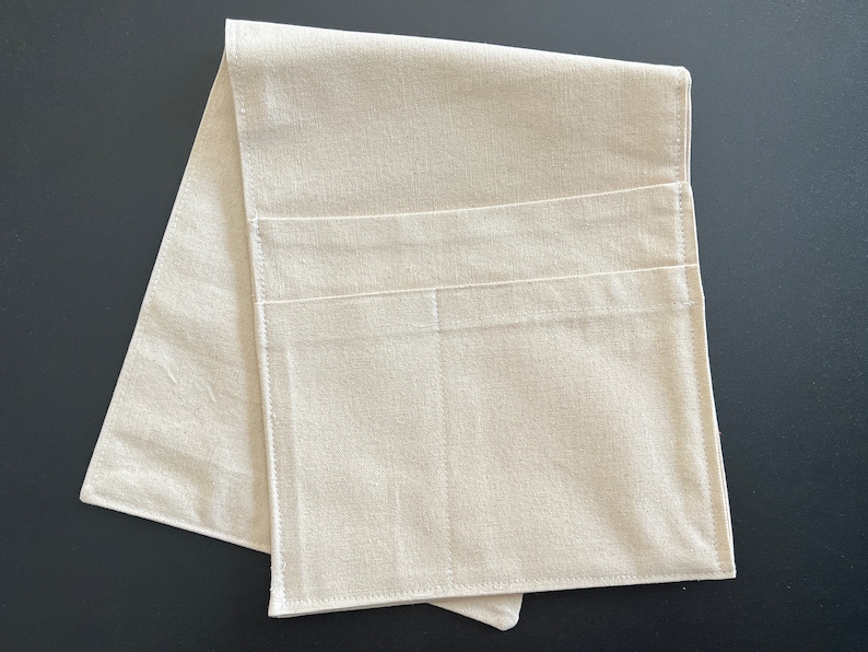 Peut inclure: Un organisateur en tissu beige clair de forme rectangulaire, dot&eacute; de deux poches horizontales cousues sur la surface avant. L'objet est compos&eacute; d'un textile &agrave; tissage simple avec des coutures visibles sur les bords et les poches, pli&eacute; &agrave; plat.