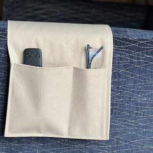 May include: A beige fabric armrest organizer featuring two rectangular pockets draped over a dark blue textured sofa arm. The left pocket contains a black remote control, and the right pocket holds a pair of eyeglasses with blue frames.