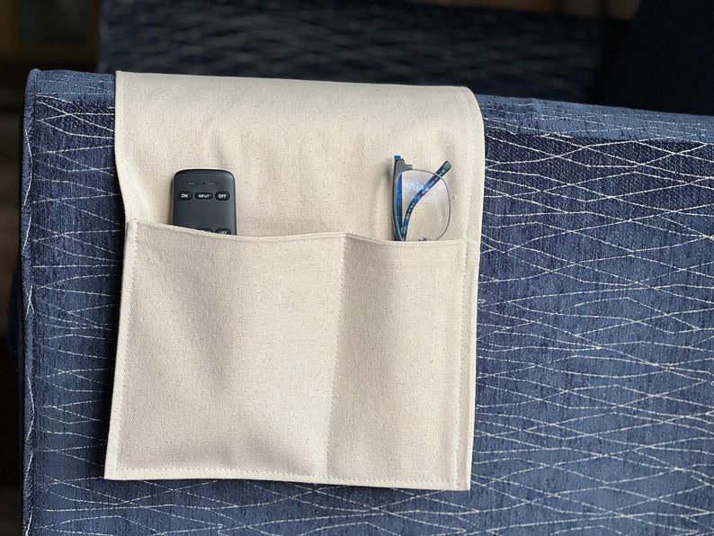 May include: A beige fabric armrest organizer featuring two rectangular pockets draped over a dark blue textured sofa arm. The left pocket contains a black remote control, and the right pocket holds a pair of eyeglasses with blue frames.