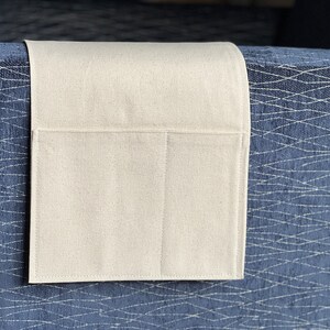 May include: A rectangular, off-white fabric armrest organizer featuring a single large pocket and a smaller side pocket, draped over the side of a dark blue upholstered furniture piece with a white, intersecting, net-like geometric pattern.