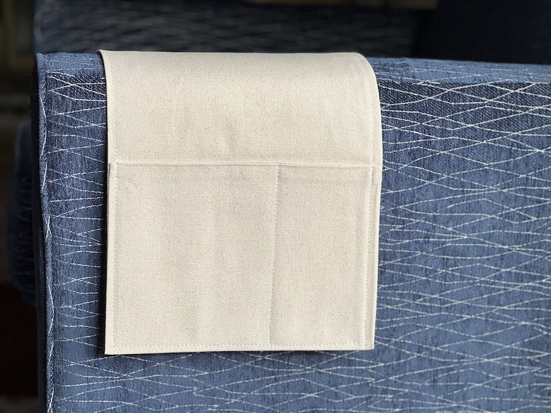 May include: A rectangular, off-white fabric armrest organizer featuring a single large pocket and a smaller side pocket, draped over the side of a dark blue upholstered furniture piece with a white, intersecting, net-like geometric pattern.