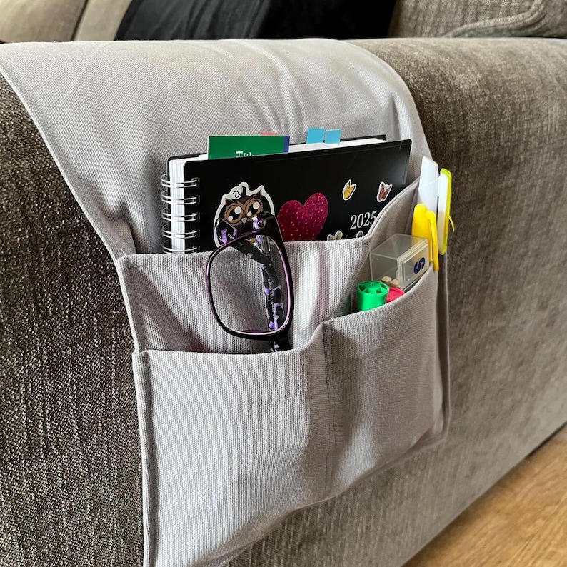Peut inclure: Un organisateur d'accoudoir de canap&eacute; en tissu gris clair avec plusieurs poches. Il contient un agenda 2025 &agrave; spirales noir avec un autocollant hibou, une paire de lunettes &agrave; monture fonc&eacute;e, un petit r&eacute;cipient en plastique et des stylos color&eacute;s.