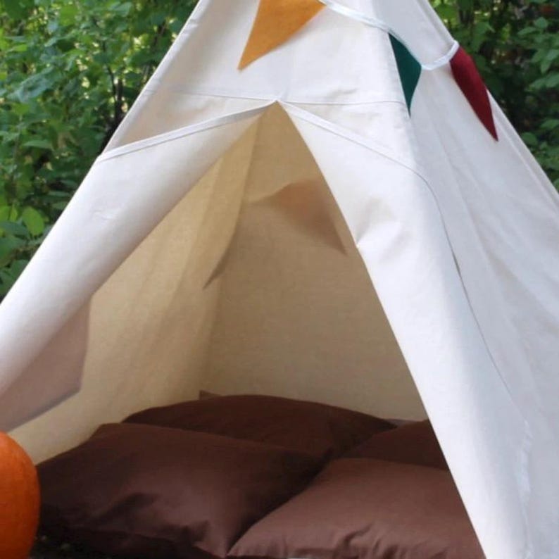 May include: A cream-colored teepee tent with brown cushions inside. The tent is decorated with a colorful banner of orange, green, and red flags. The tent is set up outdoors, surrounded by green foliage.