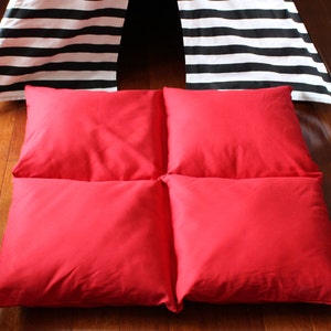 May include: Four square red cushions arranged on a wooden floor. The cushions are a solid, vibrant red. In the background, a black and white striped teepee-style structure is partially visible.