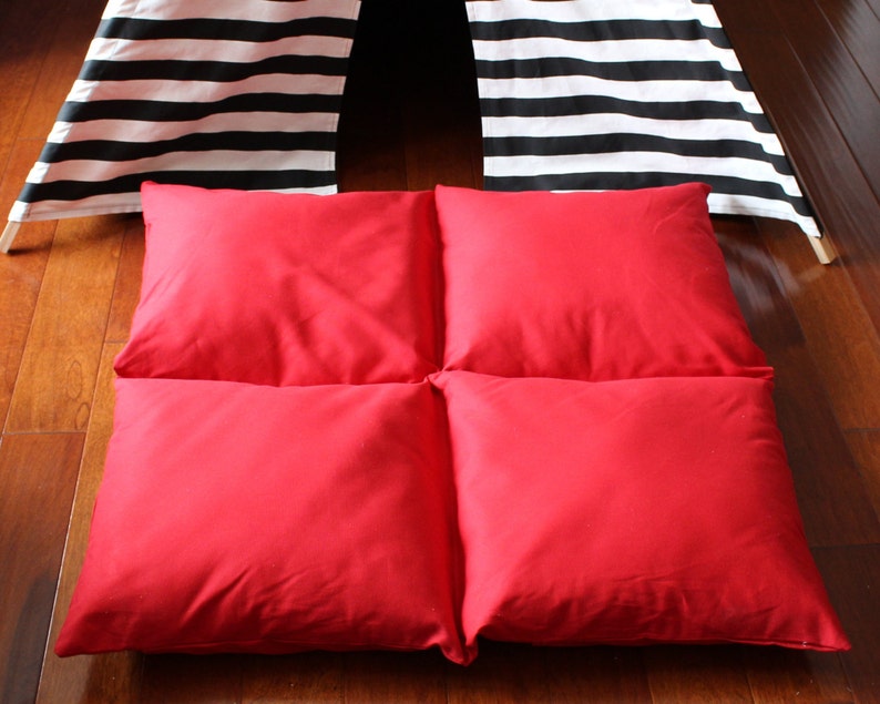May include: Four square red cushions arranged on a wooden floor. The cushions are a solid, vibrant red. In the background, a black and white striped teepee-style structure is partially visible.