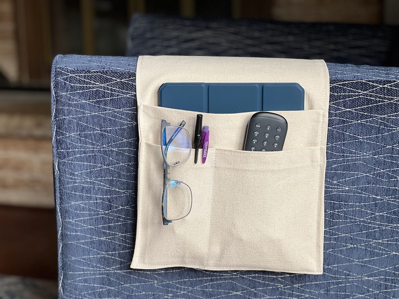 May include: A beige fabric organizer with pockets for a tablet, remote control, glasses, and pens. The organizer is attached to the arm of a blue patterned couch.