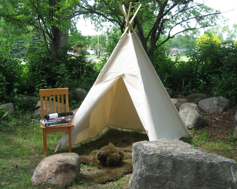 May include: A white canvas teepee tent set up outdoors with a brown stuffed animal inside. A wooden chair with a stack of books sits in front of the teepee.